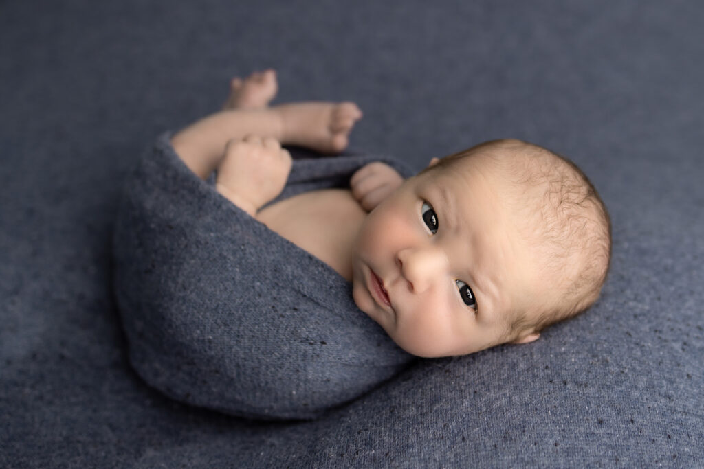 Newborn photography featuring a baby wrapped in soft blue fabric with bright eyes looking toward the camera.