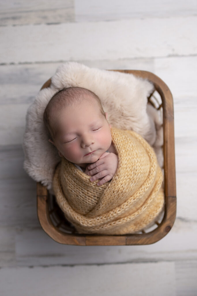 Sleeping newborn poses with hands tucked beneath the chin in a rustic wooden basket surrounded by soft beige textures.
