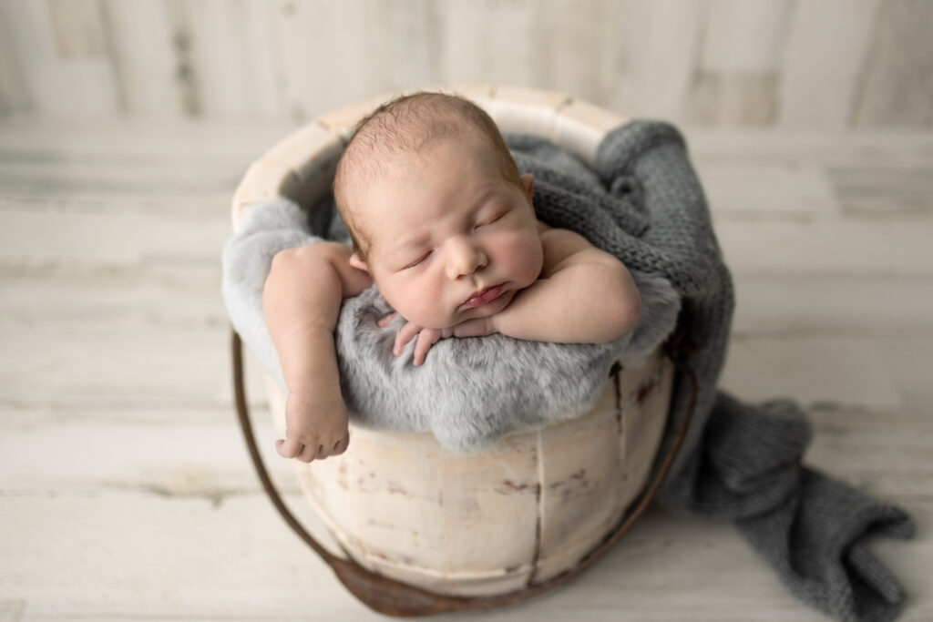 Clifton, NJ newborn photography session featuring a peacefully sleeping baby posed in a rustic wooden bucket with gray blankets.