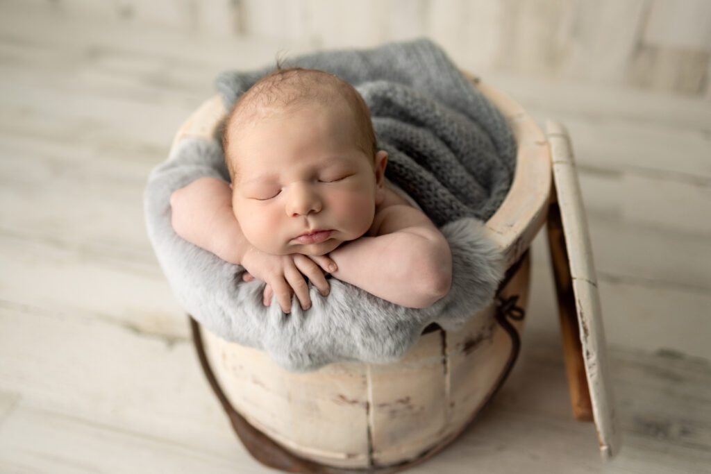 Sleeping newborn poses with hands tucked beneath the chin in a rustic wooden basket surrounded by soft gray textures.