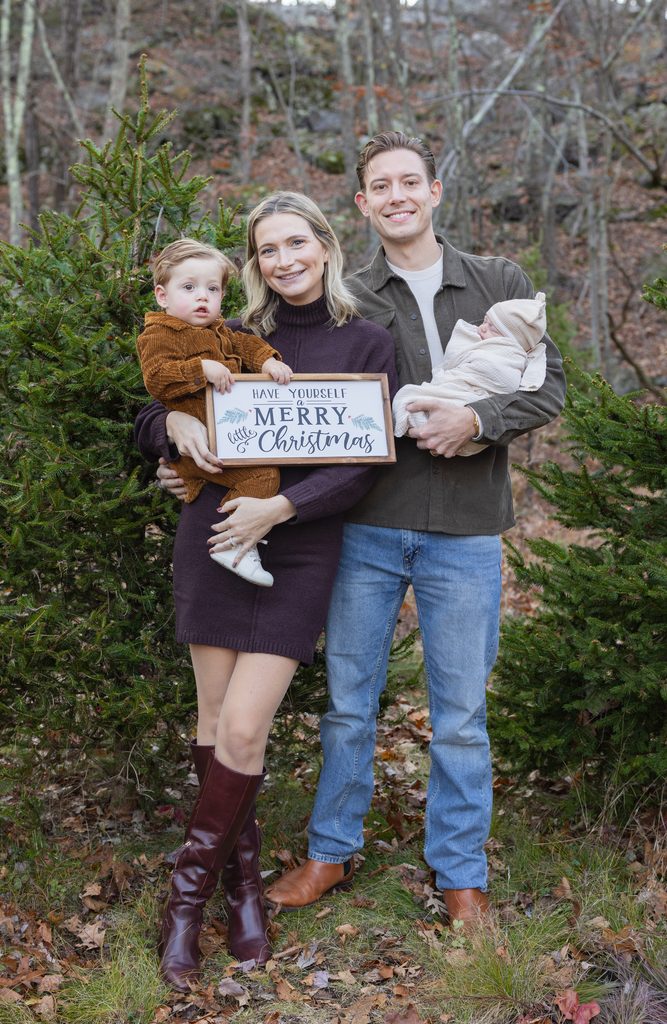 Family celebrates Christmas among evergreen trees while holding a Merry Christmas sign during a holiday mini session.
