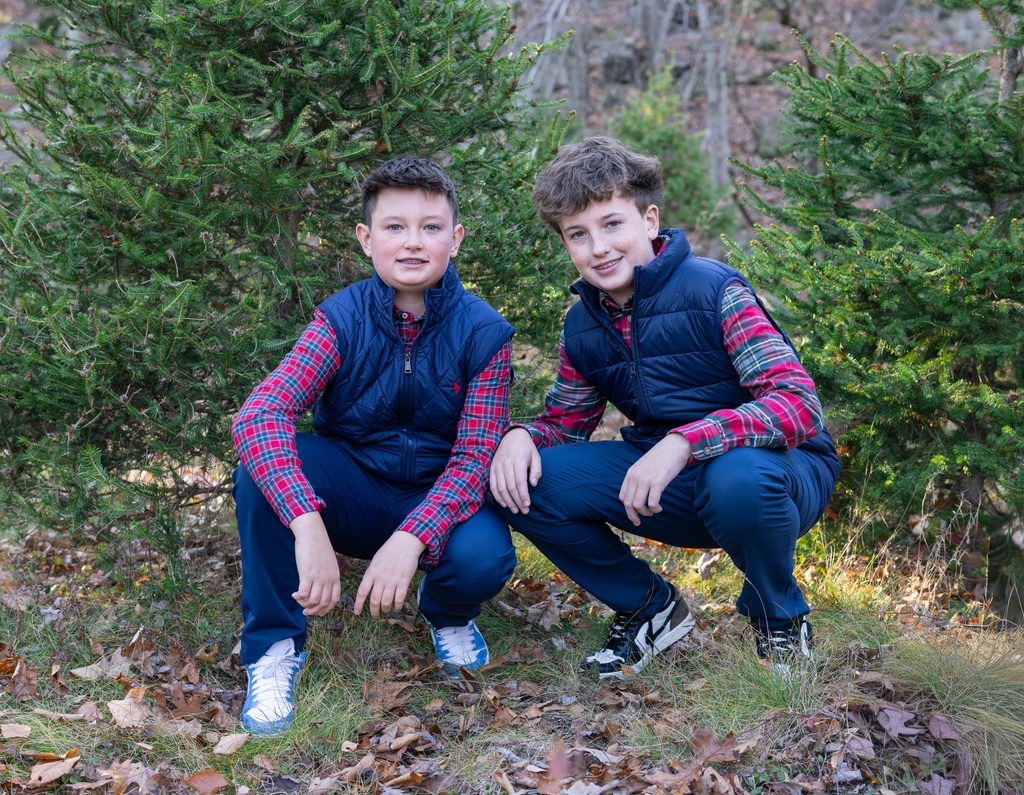 Two brothers smile together among evergreen Christmas trees during a festive holiday family photo session.
