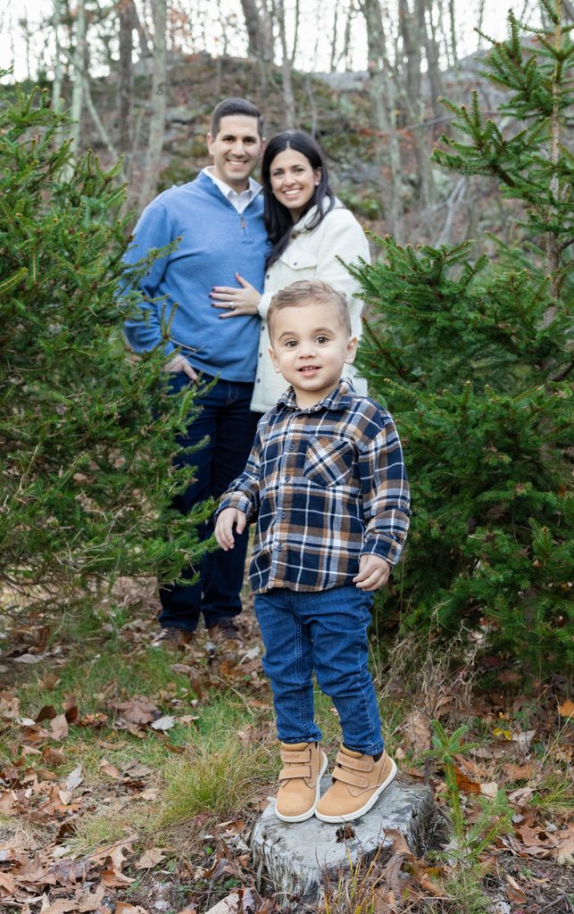 Family of three poses among Christmas trees during a relaxed holiday mini session in West Milford, New Jersey.